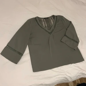 Olivgrön blus med spetsdetaljer - Snygg olivgrön blus med v-ringning och spetsdetaljer längs halsringningen och ärmsluten. Blusen har trekvartsärm och spets även i ryggen för en extra touch. Perfekt att styla med jeans eller kjol för en trendig look.