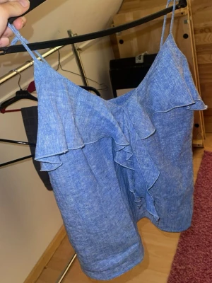 Sommar topp zara - Snygg blå linnetopp från Zara med tunna axelband och volangdetaljer framtill. Tillverkad i linne för en luftig känsla, perfekt för varma dagar. Enkel men trendig design som ger en fräsch look. Från Zara i xs