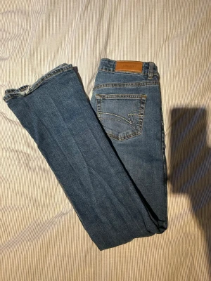 Low rise jeans från Junkyard - Säljer ett par blå low rise jeans från Junkyard med utsvängda/raka ben och snygga kontrastsömmar. Jeansen har två bakfickor med dekorativ söm och patch med logga i midjan. Perfekta till vardagslooken och tillverkade i slitstark denim. Liten slitning på benet 