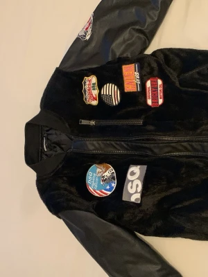 DSQUARED2 bomberjacka  - Snygg svart bomberjacka med konstläderärmar och flera färgglada patches på bröstet och ärmen. Jackan har dragkedja framtill och ribbade muddar vid ärmslut och midja. Materialet på kroppen är mjukt och lite glansigt, vilket ger en unik look. Står att det är storlek L men funkar som M också 