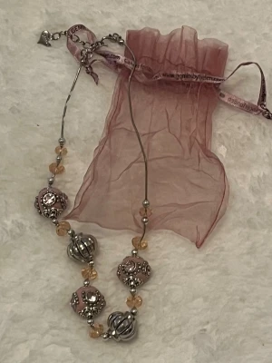 Unikt halsband med stora pärlor - Säljer ett halsband med stora dekorativa pärlor i silver och rosa, blandat med mindre glaspärlor i ljus aprikos. Kedjan är tunn och metallfärgad. Kommer med en matchande rosa organzapåse. Perfekt för dig som vill sticka ut med accessoarer!