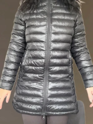 Svart lång dunjacka med dragkedja - Snygg svart lång dunjacka med quiltad design och två sidofickor med dragkedja. Jackan har en figurnära passform och går ner över höfterna. 