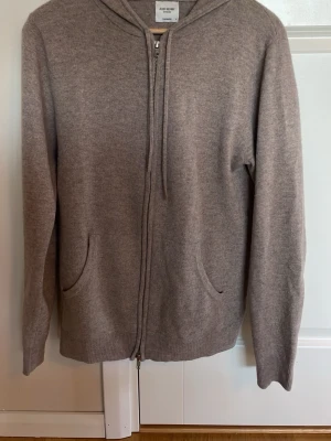 Beige hoodie från John Henric - Säljer en beige Kashmir hoodie från John Henric med dragkedja framtill och två fickor. Hoodien har en mjuk och skön känsla, ribbade muddar och justerbar huva med snören. Perfekt för dig som gillar stilrena och enkla plagg.