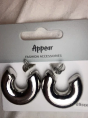 Silvriga chunky hoops från Appear - Säljer ett par chunky örhängen i silverfärgad metall från Appear. De har en rundad, tjock design och stängs med klassisk plupp. Perfekta för dig som gillar statement-smycken och vill addera lite extra bling till din look.