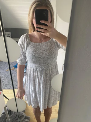 Ljus kortklänning med puffärm - Söt ljus kortklänning med smockad överdel och puffiga korta ärmar. Klänningen har en lätt utställd kjol och diskret blommigt mönster. Perfekt för sommaren! Storlek 164.