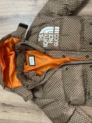 Gucci x The North Face  pufferjacka - Säljer en brun pufferjacka från Gucci x The North Face med ikoniskt monogrammönster och stor vit logga på bröstet. Jackan har orange foder, huva och stora fickor framtill. Perfekt för dig som vill sticka ut i vinter med exklusiv streetwear.