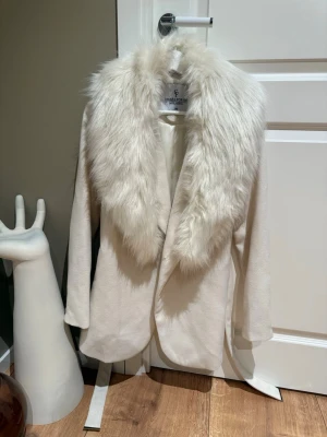 Vit kappa med fuskpäls från Chiara Forthi - Stilren vit kappa från Chiara Forthi Milano med stor, fluffig fuskpäls i vitt runt kragen. Kappan har en rak passform och släta ärmar, samt ett mjukt innerfoder för extra komfort. Perfekt för dig som vill ha en elegant och trendig look.