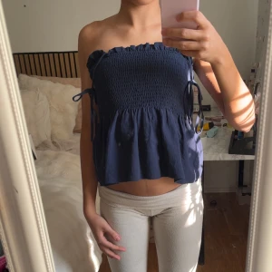 Mörkblå bandeau topp med knyt - Säljer en mörkblå bandeau topp med smockad överdel och volang nedtill. Toppen har knytbara axelband och är tillverkad i ett lätt bomullsliknande material. Perfekt för varma dagar och enkel att styla med jeans eller kjol.