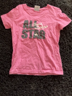 Rosa Converse All Star t-shirt XS - Cool rosa t-shirt från Converse med tryck 'All Star Chuck Taylor' i grått och vitt på bröstet. Klassisk rund hals och korta ärmar, perfekt för dig som gillar streetstyle och ikoniska märken. Materialet är mjuk bomull som känns skönt mot huden.