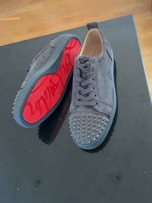 Gråa Louboutin sneakers med nitar - Säljer ett par gråa sneakers från Christian Louboutin i mocka med ikoniska röda sulor. Perfekta för dig som vill sticka ut med en lyxig och edgy stil. Låda finns tyvärr inte.