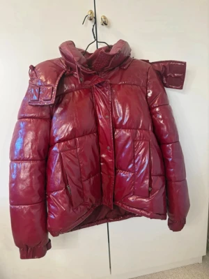 Glansig vinröd pufferjacka - Säljer en glansig vinröd pufferjacka med hög krage och stora fickor framtill. Jackan har dragkedja och tryckknappar. Perfekt för kallare dagar.