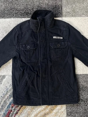 Mörkblå Fieldjacket från Hollister - Stilren mörkblå vindjacka från Hollister med hög krage och dragkedja. Två bröstfickor med knappar och två snedställda fickor framtill. Jackan har en broderad Hollister-logga på bröstet och är tillverkad i ett lätt bomullsmaterial.