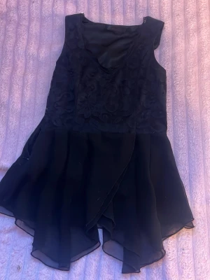 Svart omlottklänning med volang - Säljer en svart kortklänning med omlottdesign och volangkant. Klänningen är ärmlös och har ett snyggt blommigt mönster upptill. Perfekt för dig som vill ha en stilren men ändå unik look.