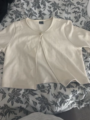 Krämvit kort kofta med knapp - Söt krämvit kofta från Gina Tricot med kortare passform och rund halsringning. Koftan har en enkel knapp framtill och korta ärmar, perfekt för lager på lager och en clean look.