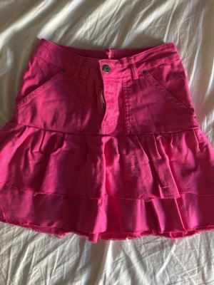 Rosa volangkjol med jeanslook - Supersnygg rosa kortkjol i jeansstil med volanger och råa kanter. Kjolen har fickor både fram och bak samt knapp och dragkedja framtill. 