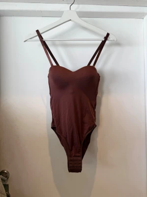 Brun body med smala axelband - Snygg brun body med smala axelband och vadderade kupor. Bodyn har en mjuk, stretchig känsla och knäppning med hyskor i grenen för enkel på- och avtagning. Perfekt att styla under skjortor eller tröjor för en clean look.