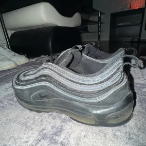 Airmax 97 - Säljer ett par svarta Nike sneakers med snörning och en stilren design. Skorna har en vågig detalj längs sidorna och en bekväm sula. Perfekta för en sportig look. 6/10 skick