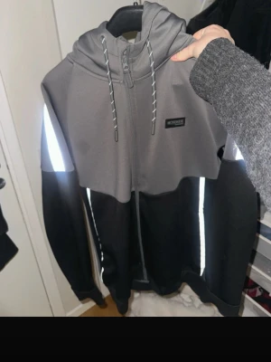 Grå och svart hoodie från McKenzie - Snygg hoodie från McKenzie. Reflekser på armar midja och snören. grå och svart med dragkedja framtill och reflekterande detaljer längs ärmarna. Huva med dragsko och logga på bröstet. Pris kan diskuteras. 
