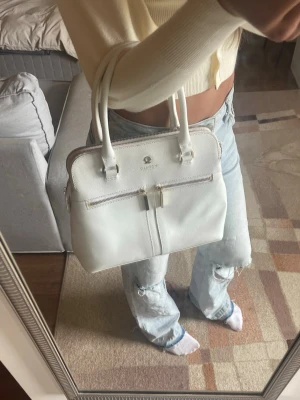 Pippa Modalu Handväska - Snygg vit designerhandväska från Modalu. Väskan har två handtag, avtagbar axelrem och guldfärgade detaljer. Tillverkad i strukturerat läder med två dragkedjefack framtill. 3 olika sektioner i handväskan varav en är fylld med flera praktiska fack. Den rymmer mycket mer än vad den ser ut att göra.  