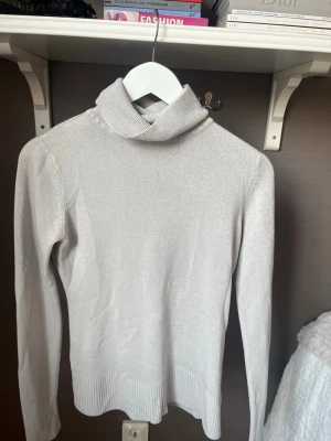 Ljusgrå polotröja i stickat - Säljer en stilren ljusgrå stickad polotröja med lång ärm och ribbade muddar vid ärmslut och nederkant. Tröjan har en hög, vikt polokrage och är tillverkad i ett mjukt, tunt material. 