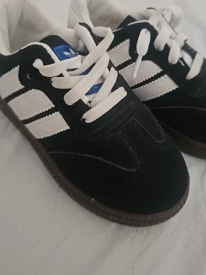 svartvita sneakers med mocka - Klassiska Adidas sneakers i svart mocka med vita läderdetaljer och tre ränder på sidan. Skorna har vita snören, blå logga på plösen och en brun gummisula med mönster. Perfekta för en sportig och stilren look.