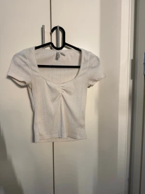 Vit ribbad topp med rynk framtill - Kortärmad vit topp med ribbad struktur och snygg rynkdetalj framtill. Djup, rundad urringning och figurnära passform. Perfekt till jeans eller kjol för en enkel men trendig look.