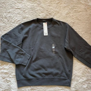 Mörkgrå sweatshirt från Uniqlo - En stilren mörkgrå sweatshirt från Uniqlo i mjuk bomullsblandning. Tröjan har rund halsringning, ribbade muddar vid ärmslut och nederkant samt en avslappnad passform. Perfekt för en casual och trendig look.