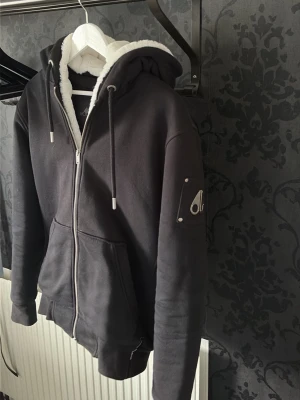 Svart zip hoodie från Moose Knuckles - Säljer en svart hoodie från Moose Knuckles med dragkedja, stor huva och vit fuskpälsfoder. Hoodien har fickor fram och den ikoniska metall-loggan på ärmen. Perfekt för dig som vill ha en stilren och varm hoodie med exklusiva detaljer.