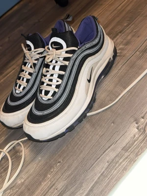 Nike Air Max 97 svart/vit sneakers - Nike Air Max 97 sneakers i svart och vitt med vågformade detaljer och reflekterande inslag. Ovandelen är i mesh och syntet, med klassisk synlig Air-sula och lila innerfoder. Ikonisk Swoosh-logga på sidan och tungan. Skön och sportig design.