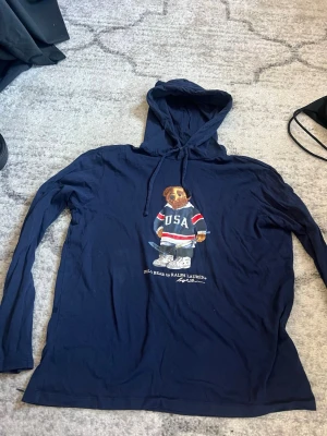Marinblå hoodie Polo Bear Ralph Lauren - Snygg marinblå hoodie från Ralph Lauren med den ikoniska Polo Bear-trycket på bröstet. Björnen har på sig en USA-tröja och håller en hockeyklubba. Hoodien har dragsko i huvan och är tillverkad i mjukt material. Perfekt för en avslappnad och sportig stil.