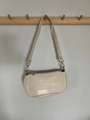 Beige axelväska Gina Tricot - Stilren beige axelväska i syntetmaterial med krokodilpräglad yta. Väskan har en avtagbar axelrem med silverfärgade metalldetaljer och dragkedja upptill. Finns ett litet fack med dragkedja i. Perfekt för dig som gillar trendiga accessoarer med lite extra attityd. I princip aldrig använd. Pris kan diskuteras!🥰