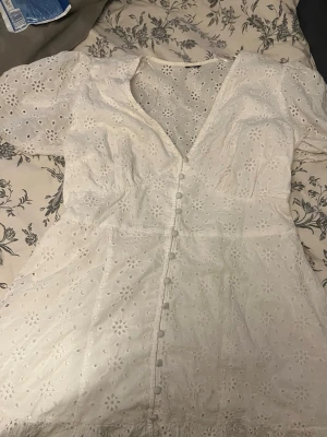 Vit kort klänning från gina - Superfin vit kort klänning i bomull med broderade blommor, puffärmar och v-ringning. Snygga klädda knappar framtill och lätt a-linjeformad passform. Perfekt för sommarens alla tillfällen! 
