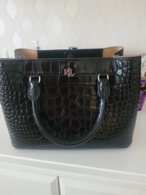 Svart handväska från Ralph Lauren - Snygg svart handväska från Ralph Lauren med RL-logga i silver framtill. Väskan har ett croco-mönstrat konstläder, två handtag och dragkedja upptill. Insidan är beige och har flera fack för smart förvaring. Perfekt accessoar för att lyfta din outfit.          Mått: Höjd 19cm. Längd 28cm. Vidd 14cm.  Handtag 10.2 cm. Kommer med dustbag. Har ej kvitto kvar men original pris är 5195kr. På zalando.   Väskan är I toppskick I princip oanvänd.