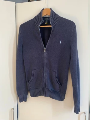Blå ziptröja från Polo Ralph Lauren - Mörkblå stickad ziptröja från Polo Ralph Lauren med ribbad krage och muddar. Tröjan har två fickor framtill och en vit broderad logga på bröstet. Kragen har en snygg detalj med ljusblå och vita ränder på insidan. Köptes på kidsbrandstore för cirka 1,5.
