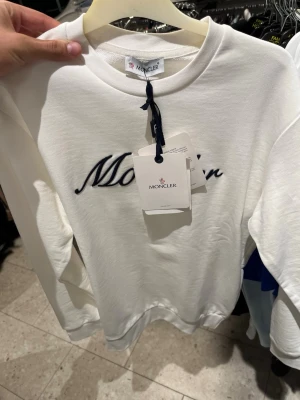 Vit sweatshirt från Moncler - Säljer en stilren vit sweatshirt från Moncler med broderad logga i svart på bröstet. Tröjan har rund halsringning och ribbade muddar vid ärmslut och nederkant. Perfekt för dig som gillar exklusiva streetwear-plagg. Äta finns olika storlekar kommer med påse och alla mer skorv privat har prislapp med den
