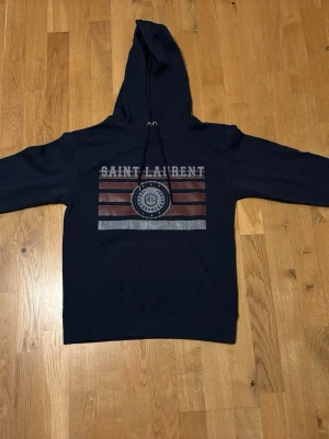 Svart Saint Laurent hoodie med tryck - Snygg svart hoodie från Saint Laurent med huva och dragsko. Framsidan har ett stort tryck med texten 'SAINT LAURENT' och grafiska ränder i rött och vitt samt ett SL-emblem. Klassisk känguruficka framtill. Perfekt för en avslappnad streetwear-look.