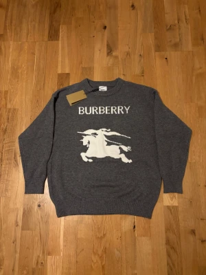 Grå stickad tröja från Burberry - Snygg grå stickad tröja från Burberry med stort vitt Burberry-tryck och riddarmotiv på bröstet. Tröjan har rund halsringning och ribbade muddar vid ärmslut och nederkant. Perfekt för dig som gillar exklusiva märken och klassisk design.