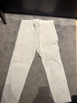 Oscar Jacobson kostymbyxor  - Snygga ljusbeige chinos med smal passform och klassisk design. Byxorna har bälteshällor, knappgylf och raka ben. Perfekta för en clean och stilren look. Tillverkade i ett mjukt bomullstyg som känns bekvämt hela dagen.