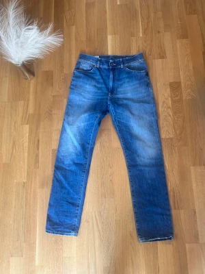 Blå raka jeans med tvättad look - Säljer ett par blå jeans med rak passform och snyggt tvättad look. Jeansen har klassisk femficksdesign, bälteshällor och dragkedjegylf. Perfekta för en avslappnad och trendig stil. Materialet är denim i bomull.