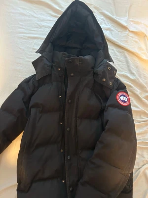 Svart dunjacka från Canada Goose - Säljer en svart dunjacka från Canada Goose med huva och klassisk logotyp på ärmen. Jackan har lång ärm, dragkedja och knappar framtill samt en puffig design som håller dig varm under kalla dagar.