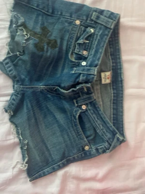 Blå jeansshorts från true religion - Säljer ett par blå jeansshorts från true religion  med råa kanter och slitningar. Shortsen har en mörk korsdetalj på ena benet och klassisk femficksdesign. Tillverkade i USA av bomull. Perfekta för en avslappnad stil!