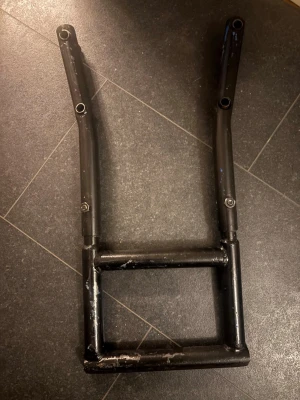 Slider tolva - Säljer en svart metallram som kan användas till cykel, moppe eller liknande projekt. Ramen har en fyrkantig nedre del och två uppstickande armar med fästhål. Perfekt för dig som gillar att bygga eller modifiera egna fordon.