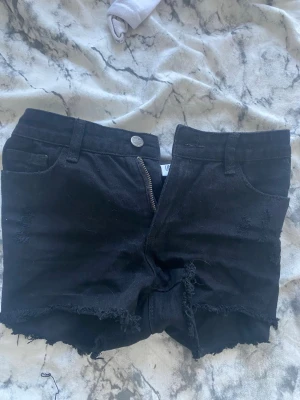 Svarta fransiga shorts med dragkedja - Svarta shorts i manchestertyg med råa, fransiga kanter och slitna detaljer. Klassisk femficksmodell med knapp och dragkedja framtill. Perfekta för en avslappnad och trendig look.