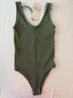 Ribbad grön body från ZARA - Snygg ribbad body i dämpad grön färg från ZARA Limitless Contour Collection. Modellen har breda axelband, rundad urringning och är tillverkad i ett stretchigt, mjukt material som sitter skönt mot kroppen. Perfekt att styla till jeans eller kjol. Aldrig använt. Storlek Xs-S. Orginalpris 169kr. 
