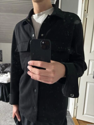 Overshirt  - Stilren svart overshirt med klassisk krage och tryckknappar framtill. Två stora bröstfickor med lock ger en cool utility-vibe. Overshirten har raka ärmar och en avslappnad passform, perfekt att bära över en t-shirt. Materialet ser ut att vara mjukt och lite tjockare.