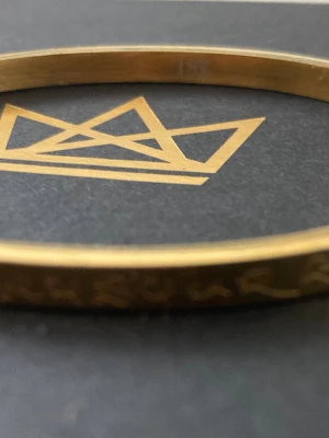 Guldfärgat armband med gravyr - Snyggt guldfärgat armband i metall med en tunn, rund form. Armbandet har en ingraverad text runt hela ytan som ger en unik och personlig touch. Perfekt för dig som gillar stilrena accessoarer med det lilla extra. Den är en thailändsk religös armband. 