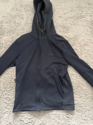 Mörkblå hoodie från BOSS - Snygg mörkblå hoodie från BOSS med dragkedja framtill och två fickor. Klassisk design med huva och dragsko. Tillverkad i mjukt material som känns skönt mot huden. Diskret BOSS-logga på bröstet för en clean look.