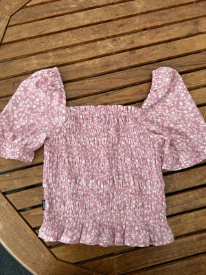 Rosa blommig topp från Seaside - Supersöt rosa topp från Seaside med vita blommor, puffiga korta ärmar och smockad kropp. Toppen har volangkant nedtill och är perfekt för dig som gillar romantisk och trendig stil. Tillverkad i mjuk bomull som känns skön mot huden.