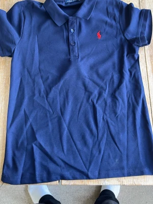 Marinblå pikétröja från Ralph Lauren - Klassisk marinblå pikétröja från Ralph Lauren med korta ärmar och krage. Tröjan har knappar framtill och den ikoniska röda logotypen broderad på bröstet. Tillverkad i mjuk bomull som känns skön mot huden.