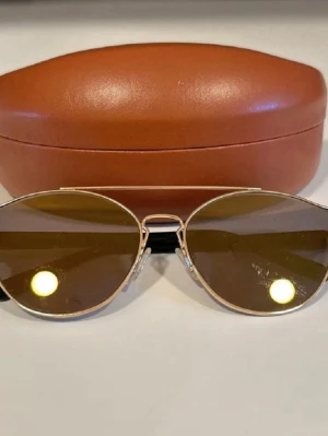Missoni pilotbågar solglasögon guld - Snygga solglasögon från Missoni med klassisk pilotform. Bågarna är i guldigt metall och glasen har en mörk ton. Svarta detaljer på skalmarna där Missoni-loggan syns. Kommer med ett brunt hårt fodral med guldfärgad logga.